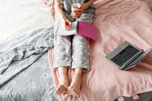 Kobieta w piżamie, siedzi na łóżku i notuje coś z kubkiem herbaty w ręce. Obok niej laptop.