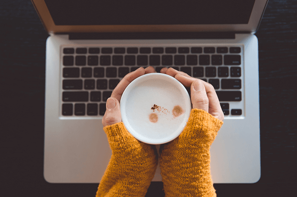Widok z góry na dłonie trzymające filiżankę z kawą ponad klawiaturą laptopa.