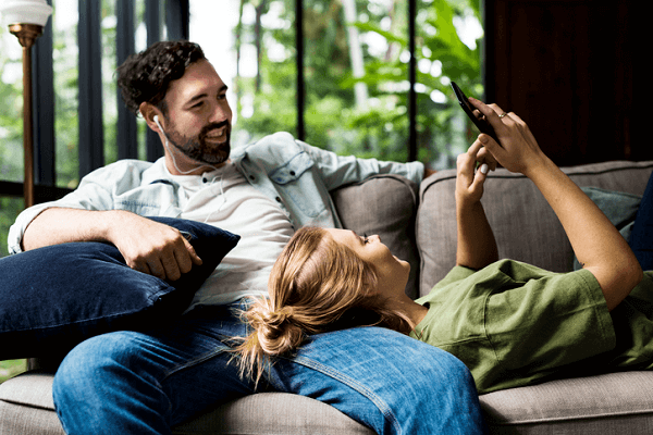 Para uczy się wspólnie na kanapie w domu. Mężczyzna ma założone słuchawki, kobieta korzysta ze smartfona.